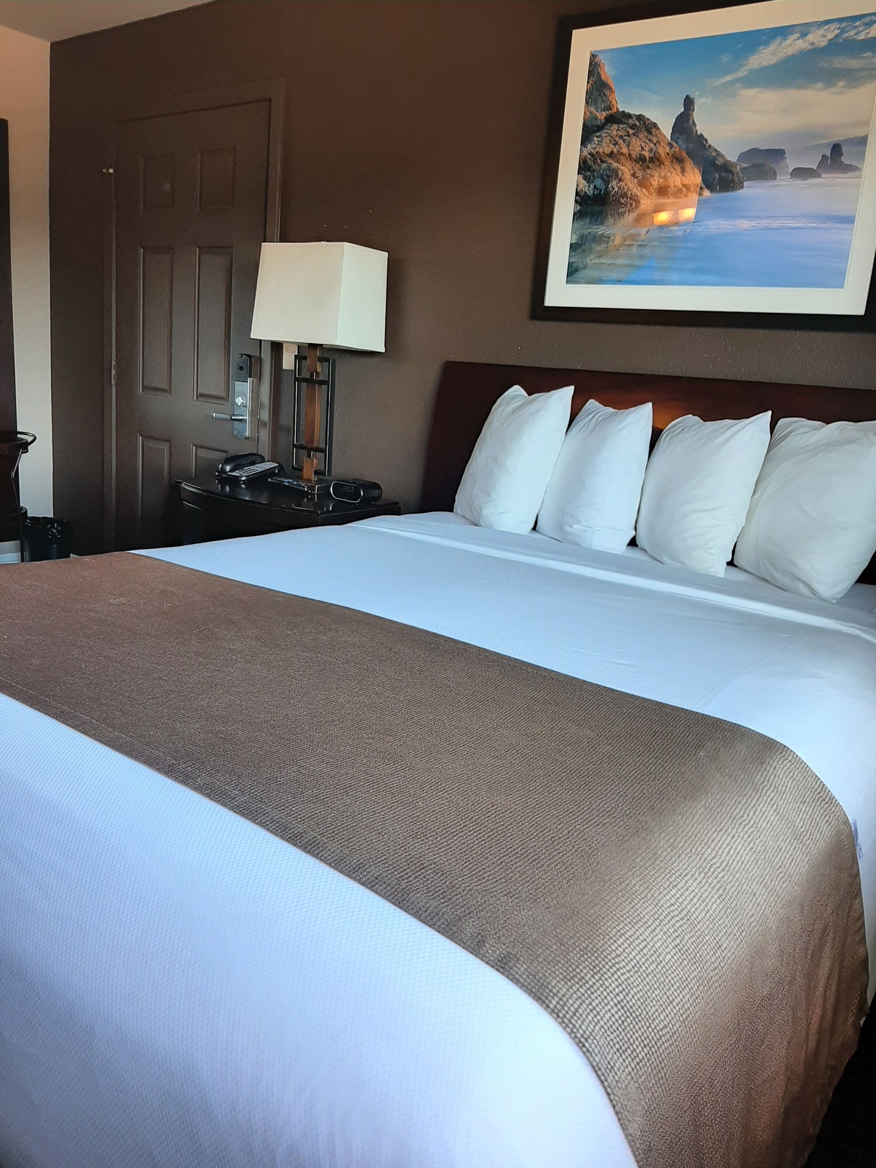 A neatly made bed with white bedding and a brown throw blanket, a bedside lamp, a telephone, and a framed picture of a coastal landscape on the wall.