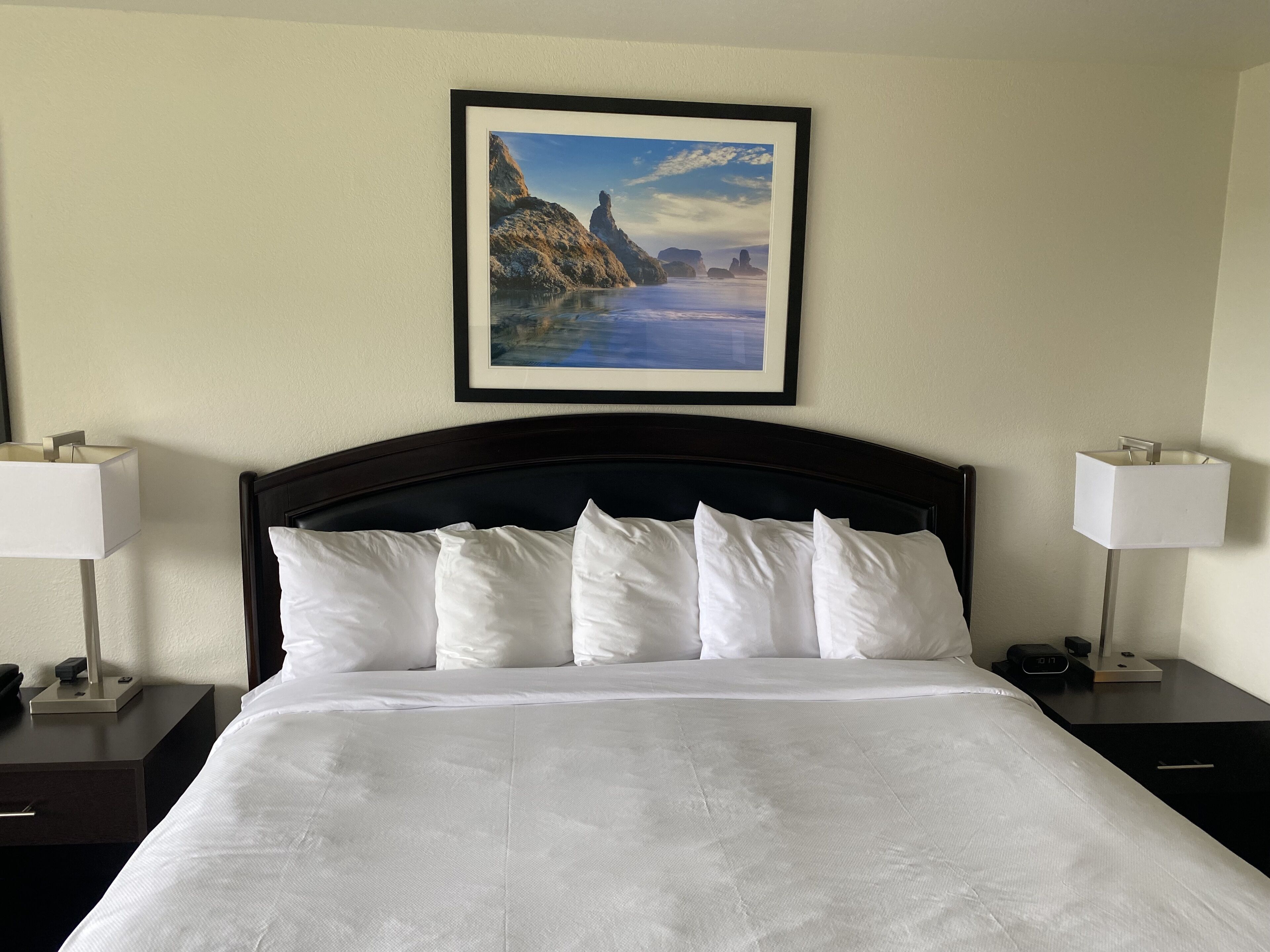 A neatly made bed with white linens, a framed landscape painting above it, and bedside lamps on either side.