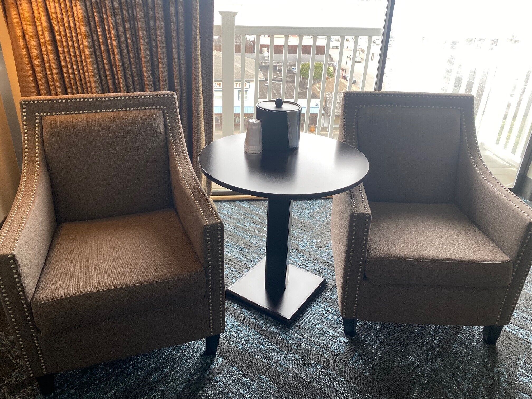 Two armchairs with a round table in between, a window with a view of a fence and buildings, and a patterned carpet.