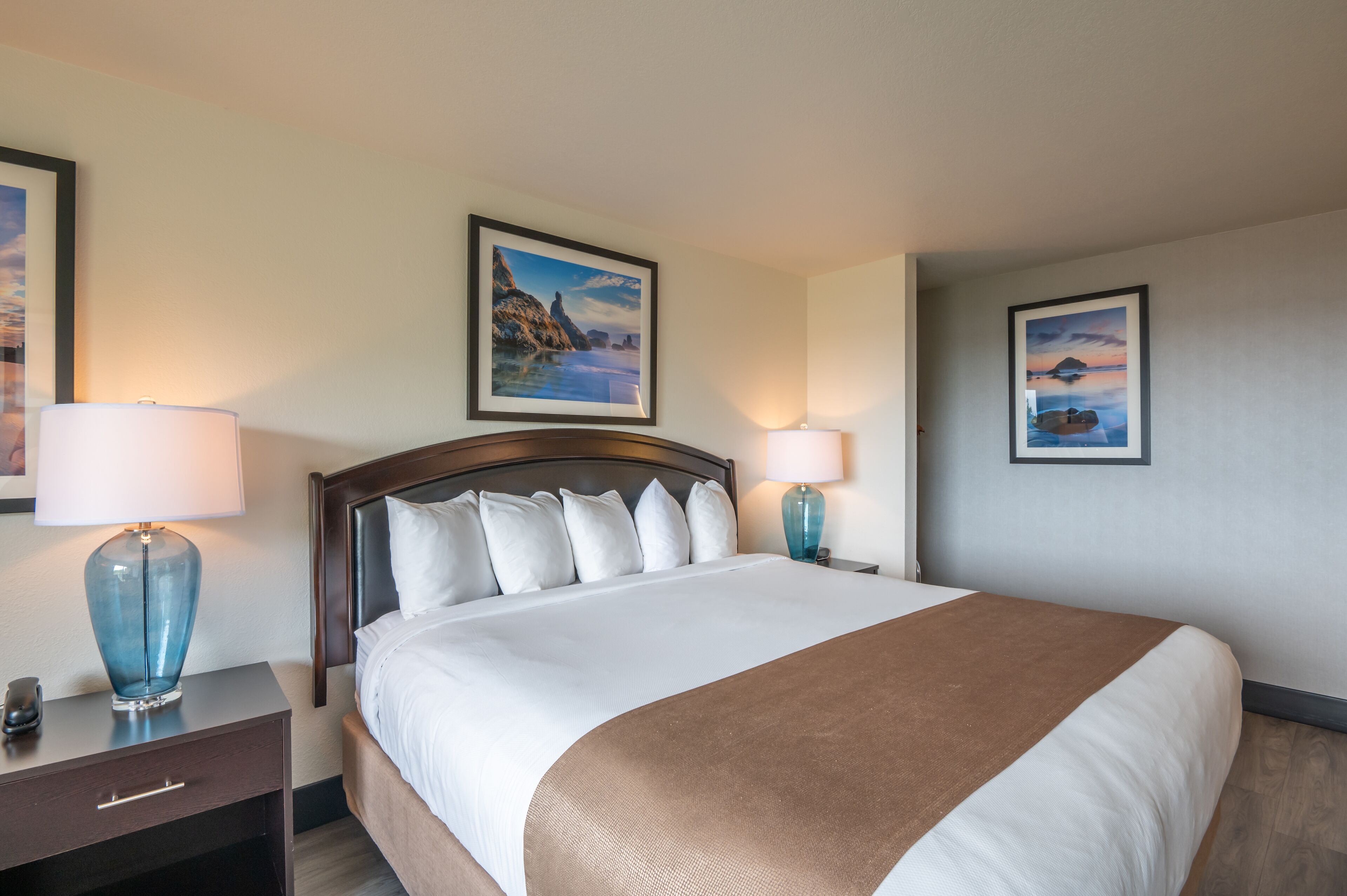 A neatly made bed with white and brown bedding, two bedside lamps, a nightstand with a drawer, and two framed pictures on the wall.