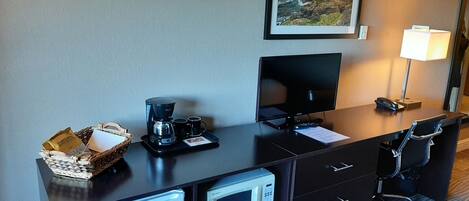 Habitación de hotel con escritorio, cafetera, monitor, lámpara, microondas, cesta con una tarjeta y un cuadro en la pared.