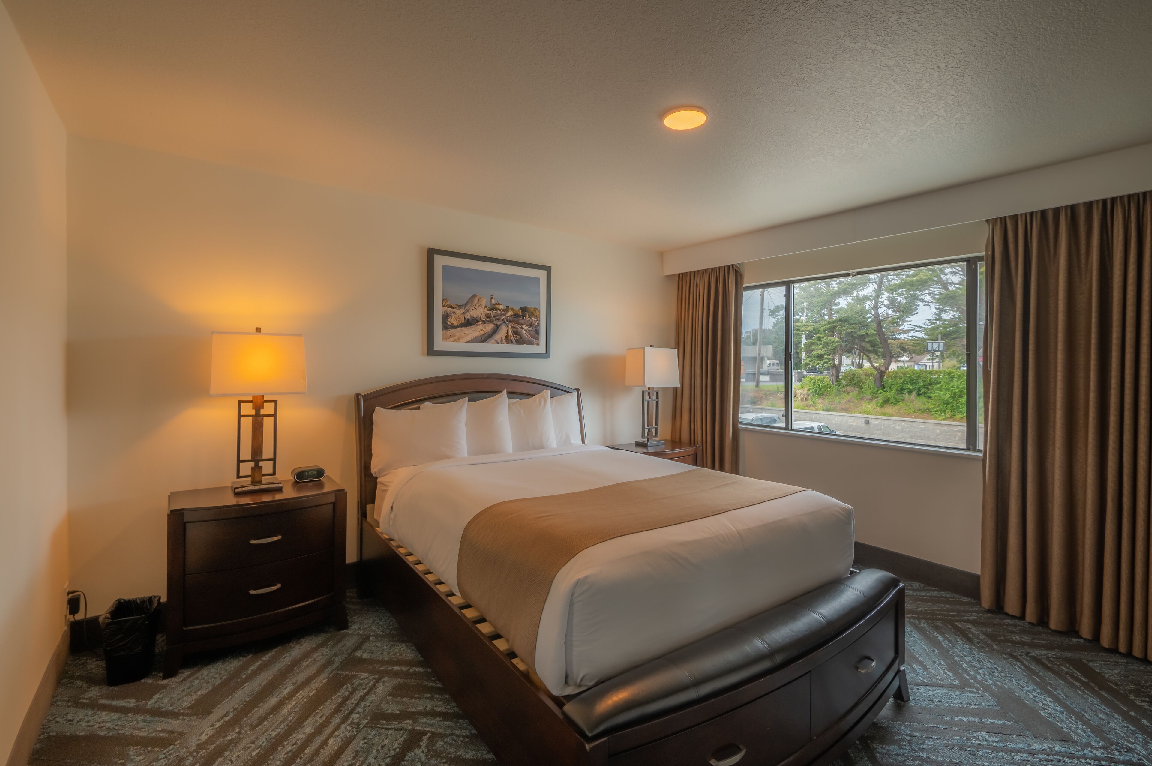 A hotel room with a large bed, two bedside tables with lamps, a window with curtains, and a painting on the wall.