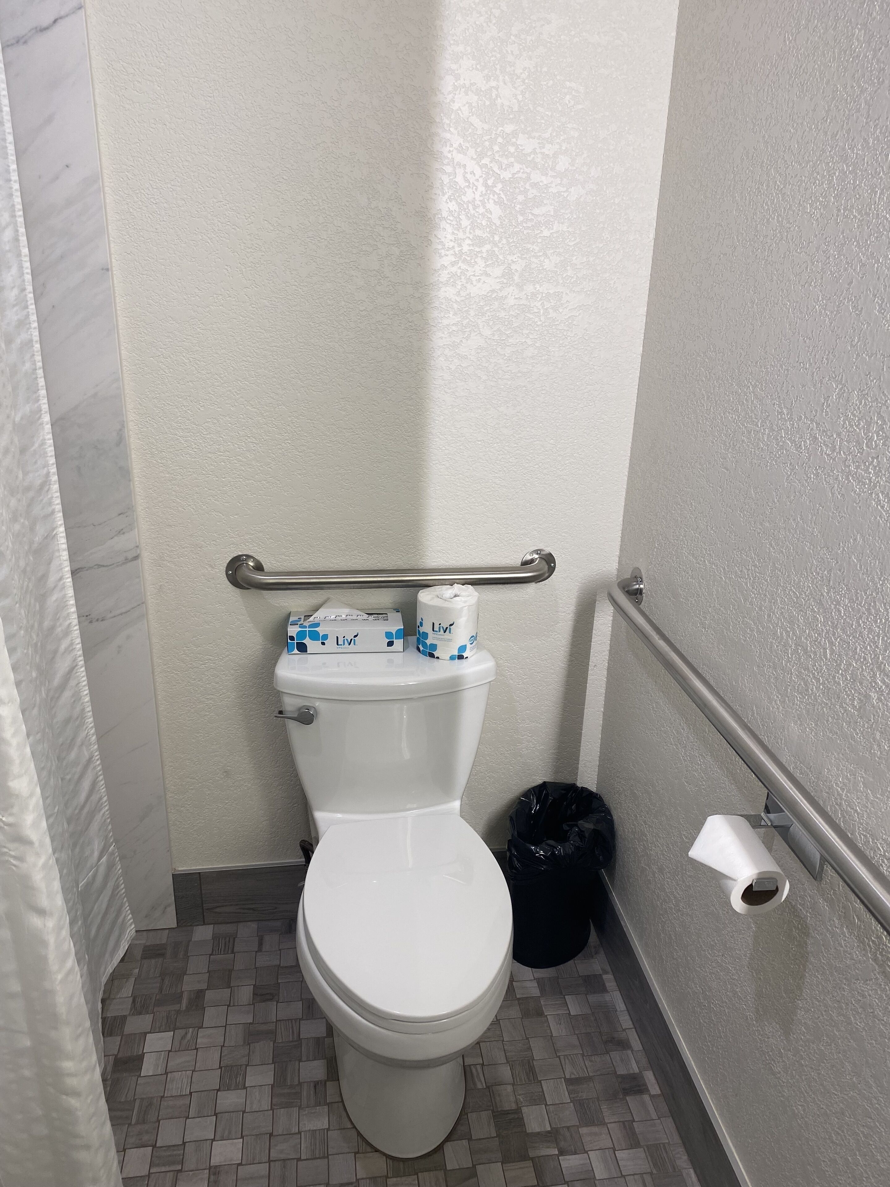 A small, compact bathroom with a white toilet, a handrail, and a trash bin.