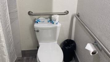 A small, compact bathroom with a white toilet, a handrail, and a trash bin.