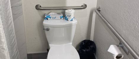 A small, compact bathroom with a white toilet, a handrail, and a trash bin.