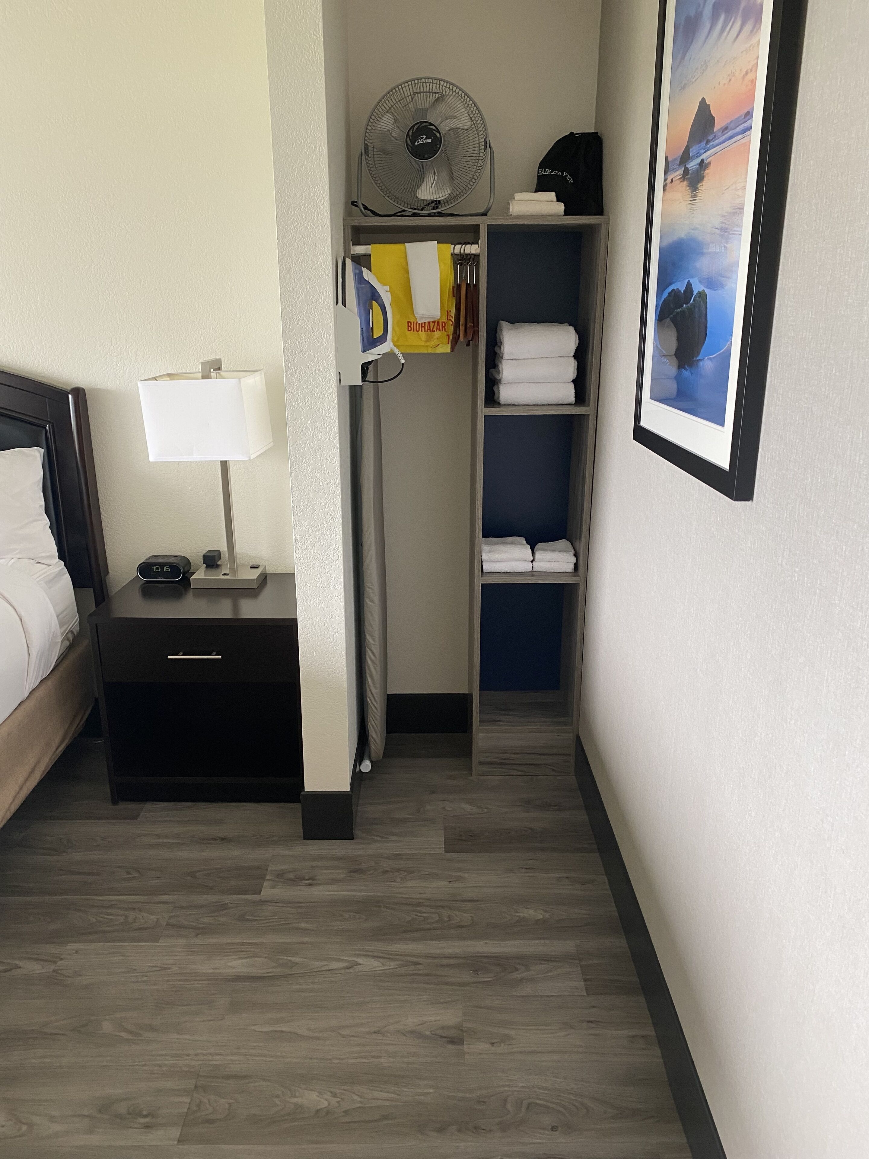 Une chambre d’hôtel comprenant un lit, une table de chevet, une lampe, une commode, un placard avec des serviettes pliées, un ventilateur et un tableau encadré au mur.