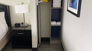 Une chambre d’hôtel comprenant un lit, une table de chevet, une lampe, une commode, un placard avec des serviettes pliées, un ventilateur et un tableau encadré au mur.