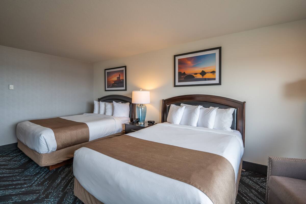 A hotel room with two beds, a nightstand with a lamp, a chair, and two framed pictures on the wall.