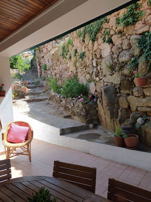 Une terrasse extérieure couverte, avec des murs en pierre, des marches en terrasses, des plantes en pot et des meubles en bois.