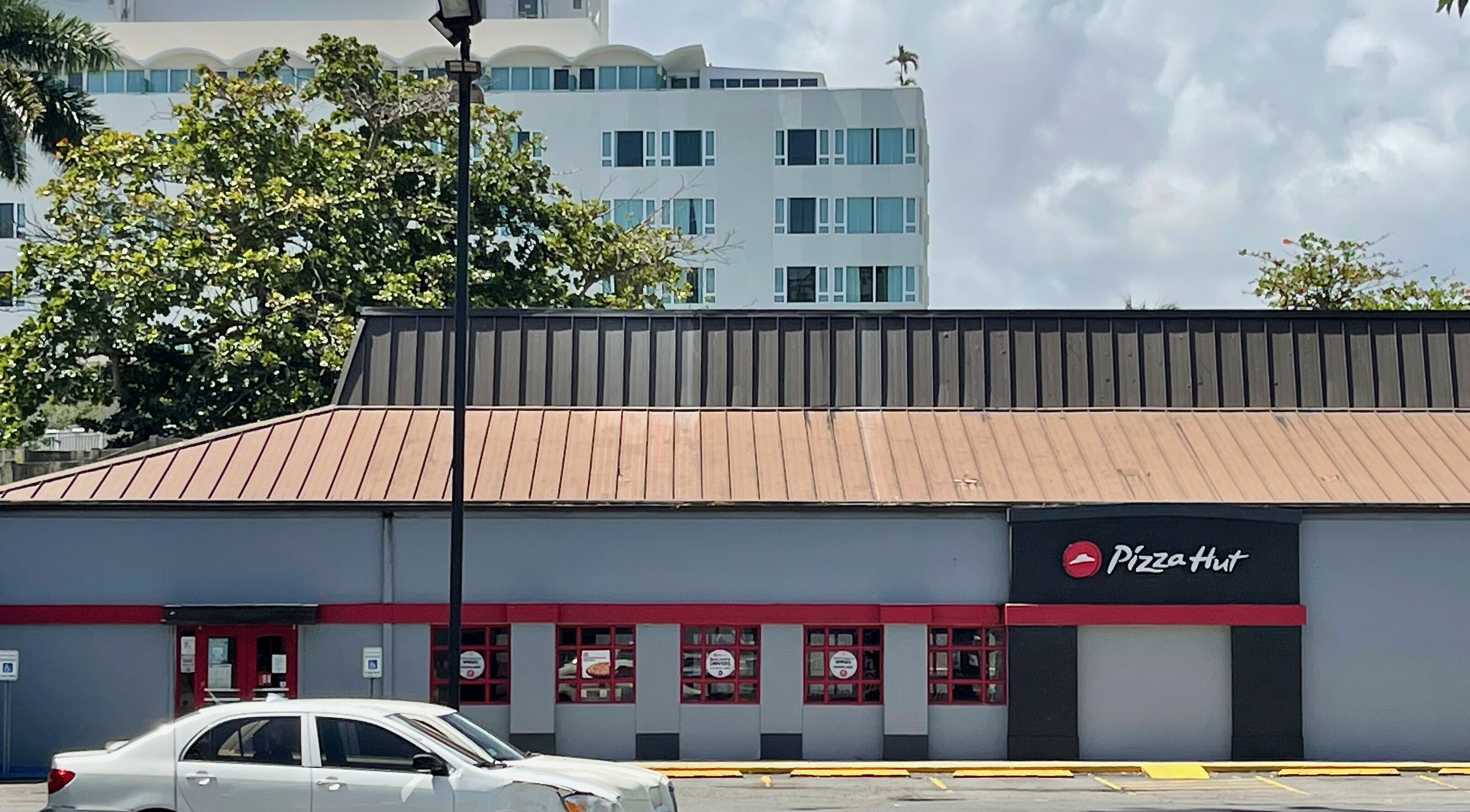 Un restaurant Pizza Hut avec une façade rouge et grise, une voiture garée devant et un bâtiment au design similaire en arrière-plan.