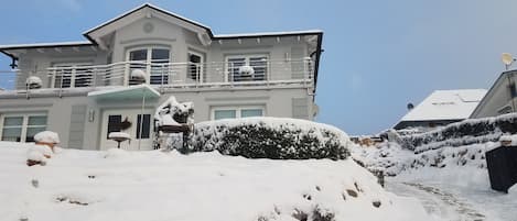 A two-story house with a balcony, surrounded by snow-covered ground and bushes.