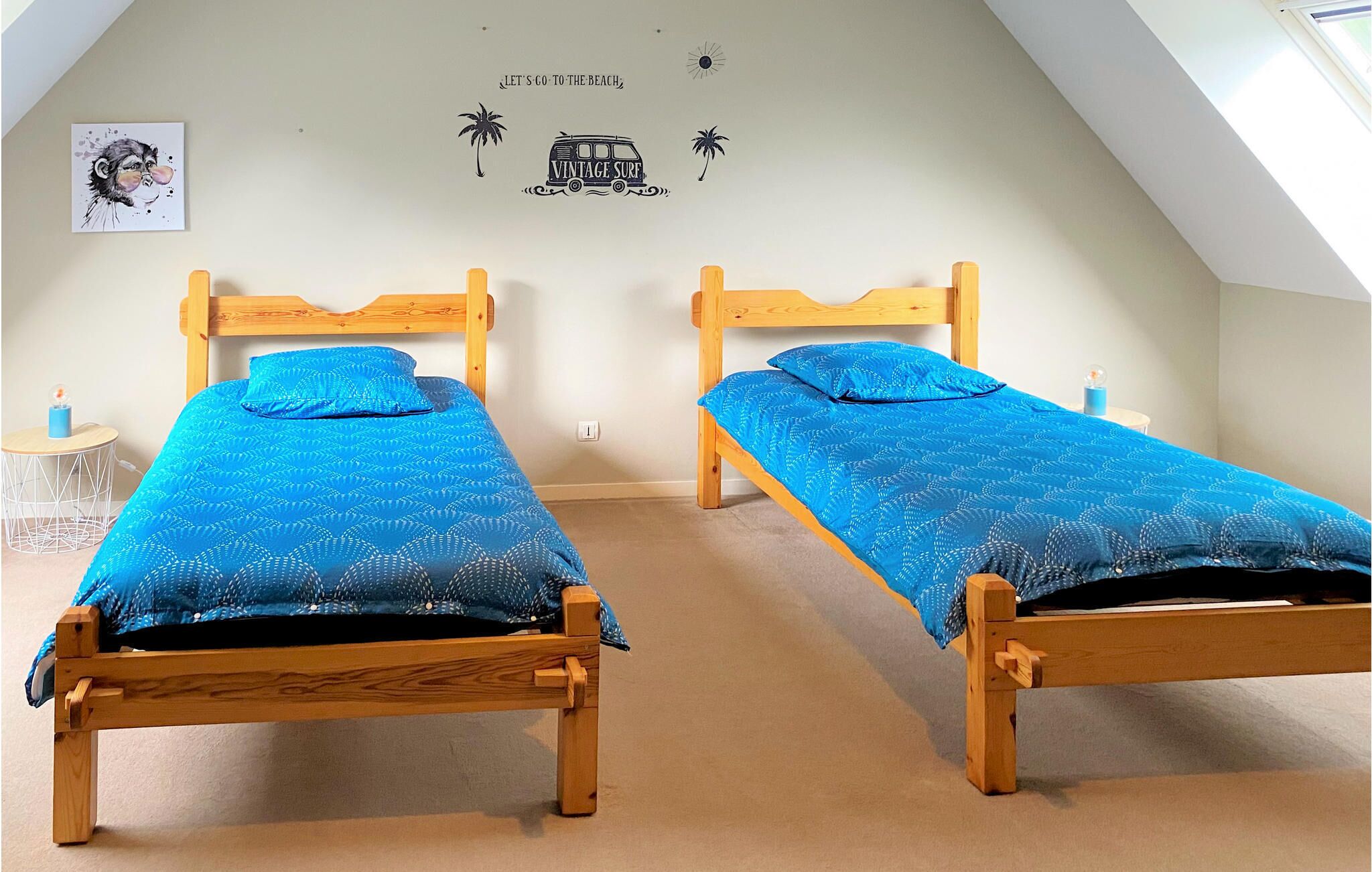 Une pièce avec deux lits en bois, chacun recouvert d’une courtepointe à motifs bleus. Un mur est décoré d’un autocollant représentant un vieux van et des palmiers, et une petite table ronde porte une bougie.