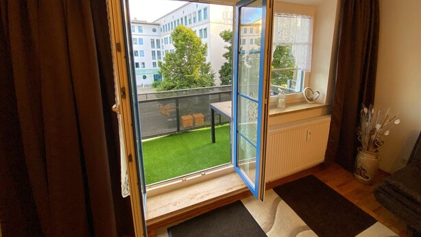 A room with a sliding glass door leading to a balcony with a view of buildings and a tree.
