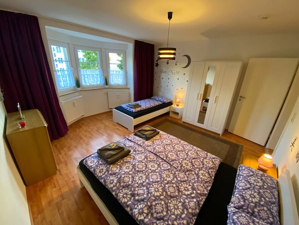 A bedroom with two beds, a wardrobe, a bedside table with a lamp, and a window with curtains.