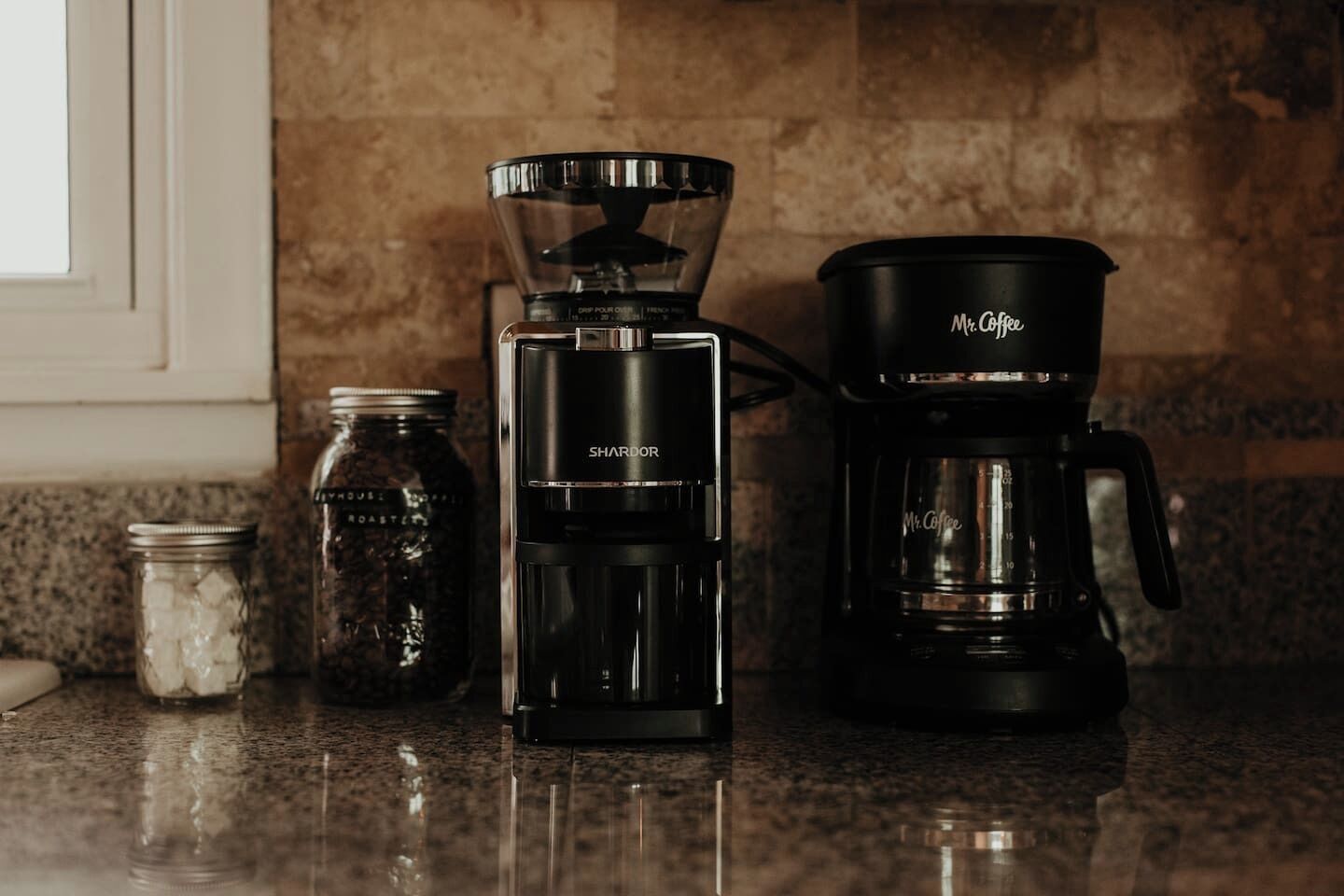 Een koffiezetapparaat, een koffiemolen en een pot koffiebonen op een aanrecht.