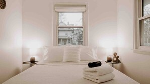 A minimalist bedroom with a large bed, two bedside tables, and a window with a view of a house. - CITY COTTAGE, CITY COTTAGE GETWAY, FENCE BACKYARD (Saint Paul)