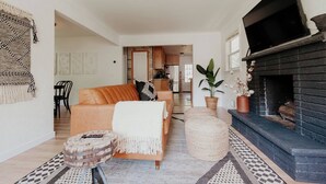 A living room with a sofa, ottoman, and a fireplace. There is a kitchen in the background. - CITY COTTAGE, CITY COTTAGE GETWAY, FENCE BACKYARD (Saint Paul)