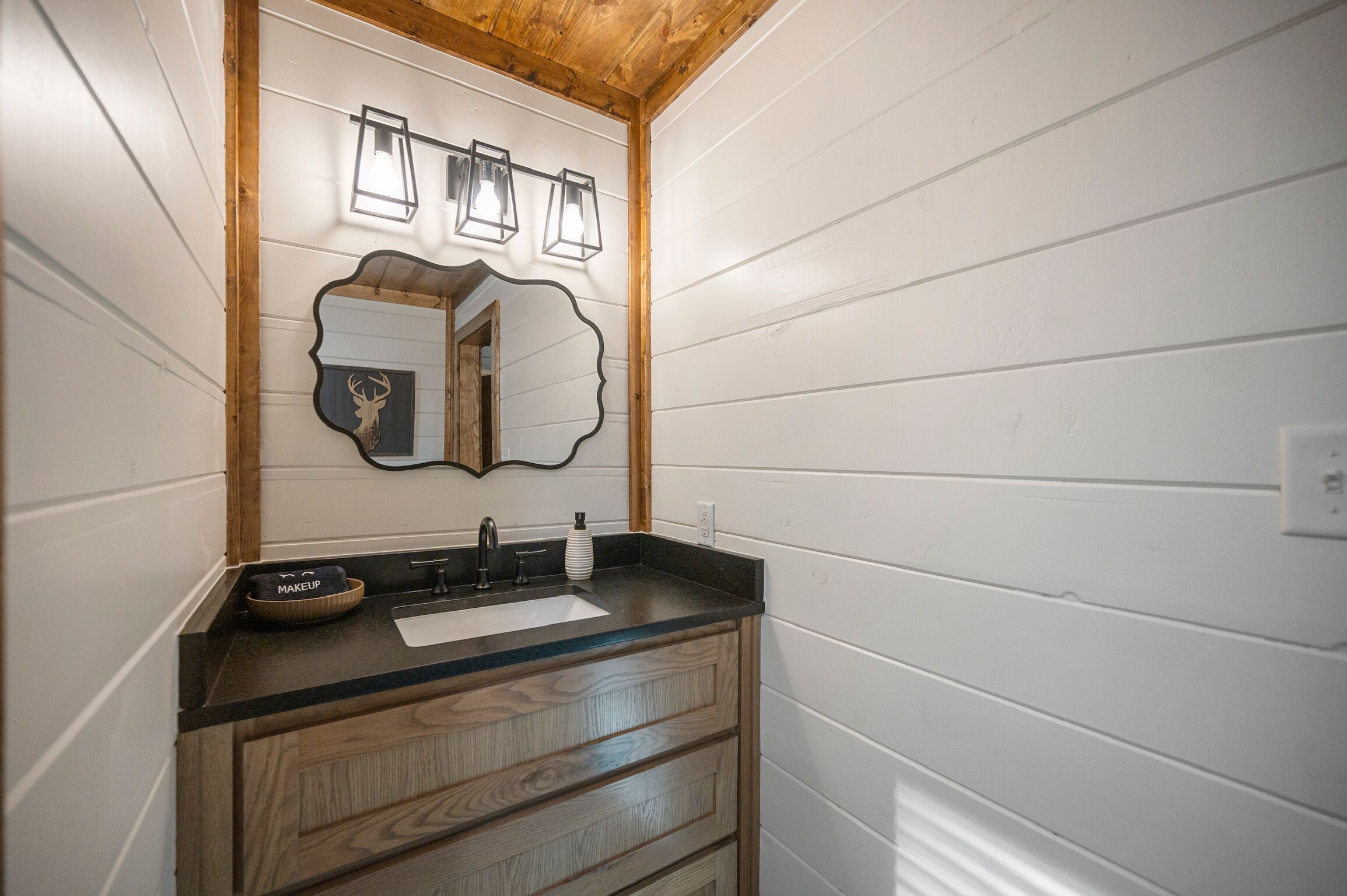 Un baño con tocador de madera, un lavabo y un espejo con decoración de cabeza de ciervo. Hay tres luces fijadas a la pared sobre el espejo.