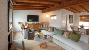 Una sala de estar acogedora con techo de madera alto, un sofá blanco grande, un sillón gris y una mesa de centro redonda.