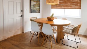 Un comedor con una mesa redonda de madera, sillas blancas, una lámpara colgante de mimbre y una ventana con persianas.