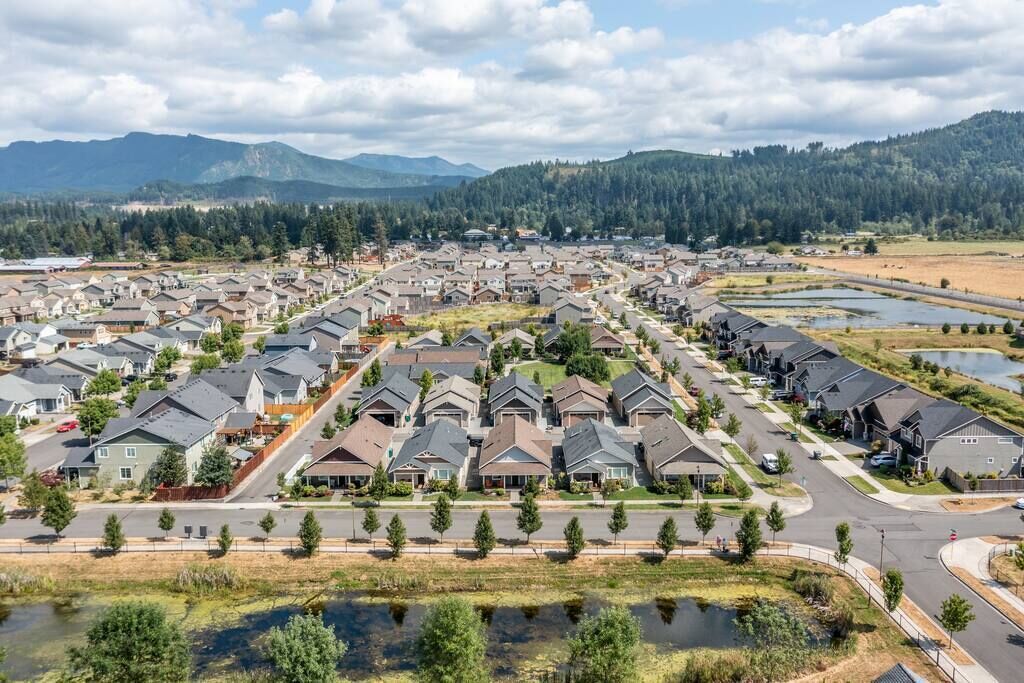 A residential area with houses arranged in rows along a main road, surrounded by greenery and a lake.
