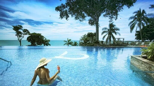 Una piscina con una persona sentada en el agua, rodeada de palmeras y un cielo despejado.