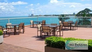 Un patio al aire libre con mesas y sillas, con vista a un cuerpo de agua y un paisaje urbano.