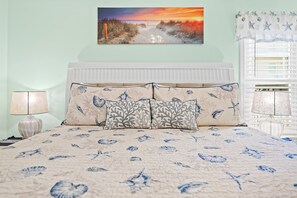 A bedroom with a bed featuring a sea turtle and starfish pattern, a nightstand with a lamp, and a painting of a sunset beach scene. - "SALT THERAPY"  Waterfront Condo. Destin, Florida (Destin)