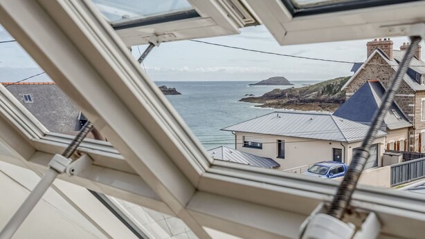 Blick durch ein Dachfenster auf ein Küstengebäude mit einer Stein-Fassade, einem blauen Dach und Blick auf das Meer sowie eine entfernte Insel.