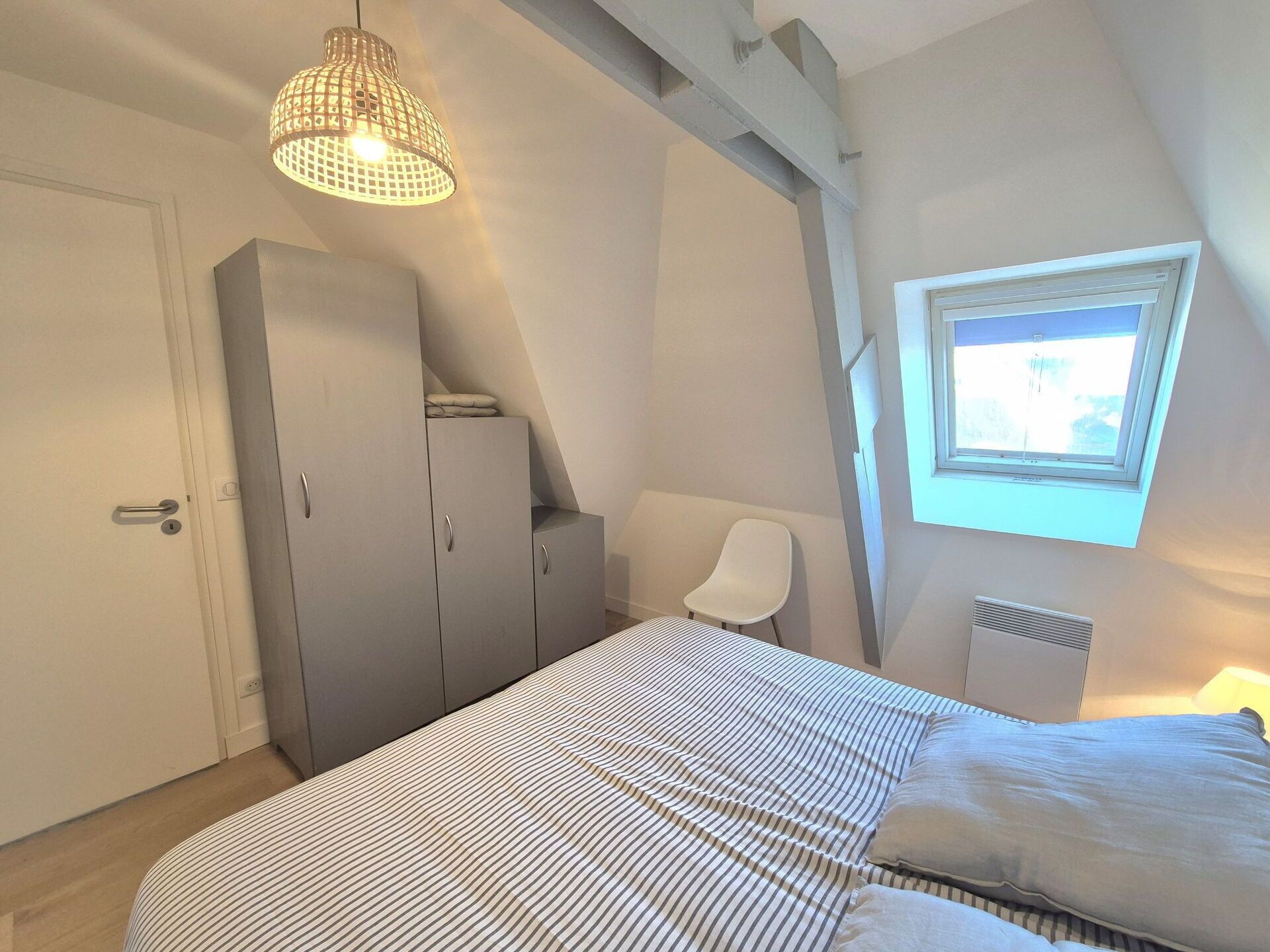 Un dormitorio con cama, armario, silla, ventana y lámpara colgante.