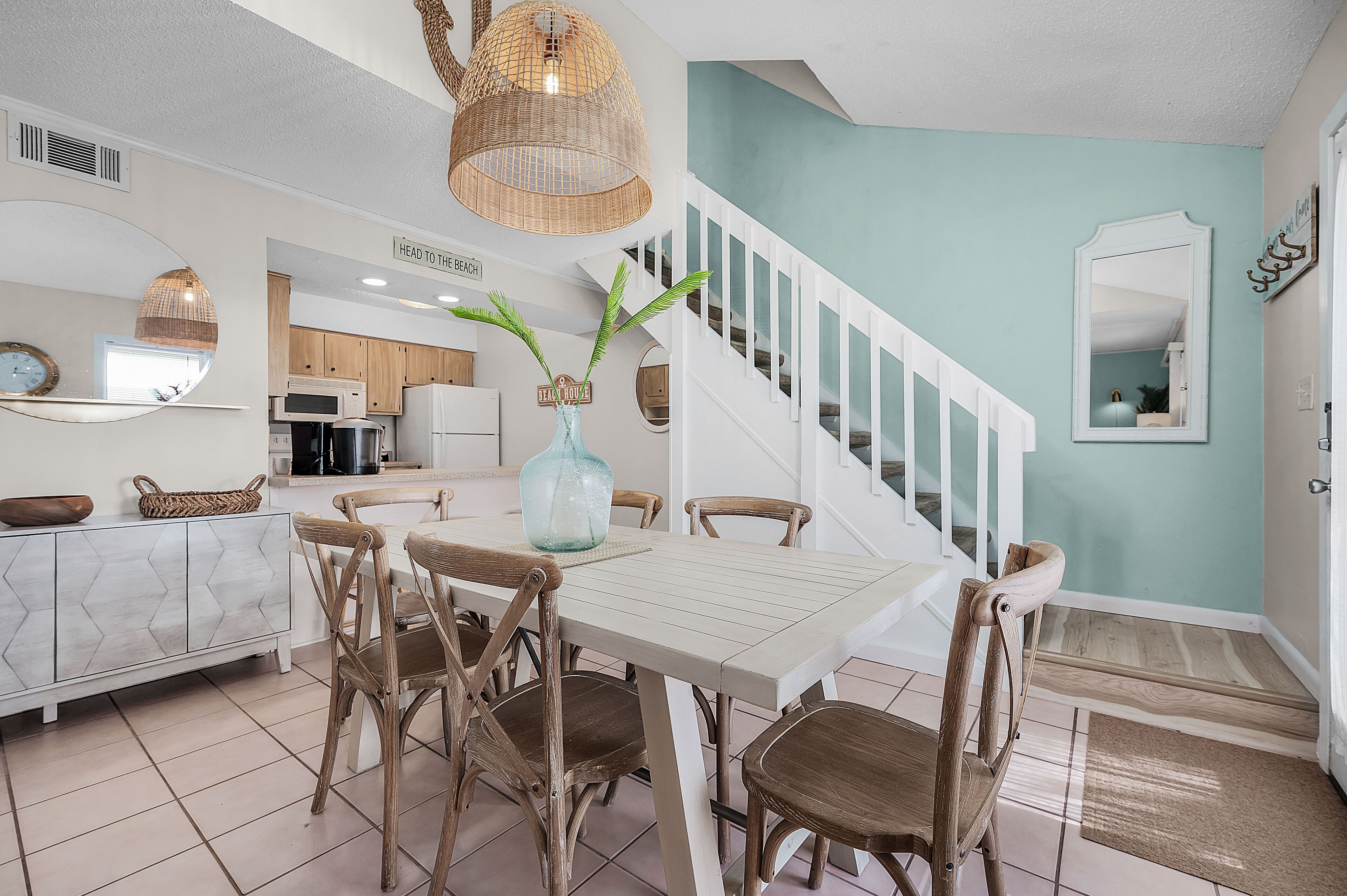 A dining area with a white table and wooden chairs, a kitchen in the background, and a staircase with white railings.