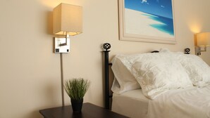 Una camera d'albergo con un letto, un comodino con una lampada, una pianta in vaso e un quadro raffigurante una spiaggia.