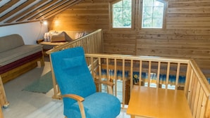 A cozy attic bedroom with a wooden bunk bed, a blue chair, a small table, and a window overlooking greenery.