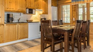 A wooden cabin-style kitchen with a dining area, featuring a large table and chairs, wooden cabinets, a microwave, and a refrigerator.