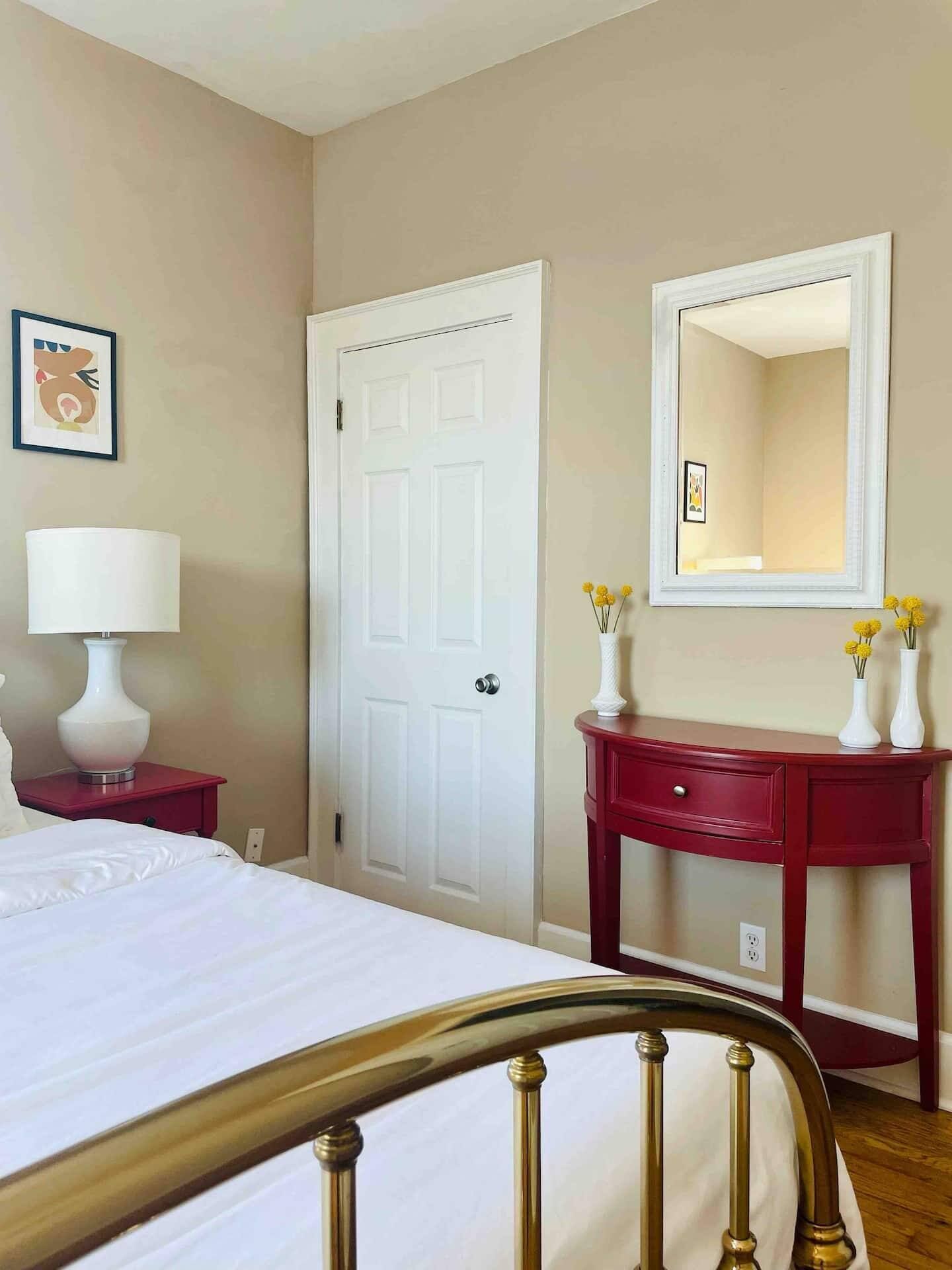 Una habitación de hotel con una cama, una mesita de noche con lámpara, un aparador con jarrones, una puerta y un espejo.