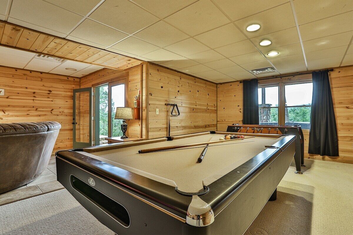 A pool table in a room with wooden walls, a sofa, and a window.
