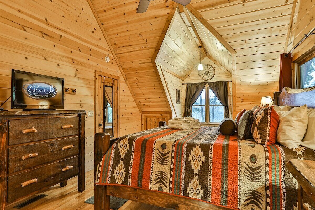 Une cabane avec un intérieur en bois, un lit recouvert d’une couverture à motifs, une commode et un téléviseur qui affiche le logo de « Blue Sky Cabin Rentals ».