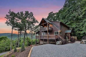 A two-story log cabin with a balcony, surrounded by trees and a gravel driveway.