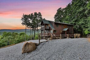 A cabin with a balcony, surrounded by trees and a gravel driveway.