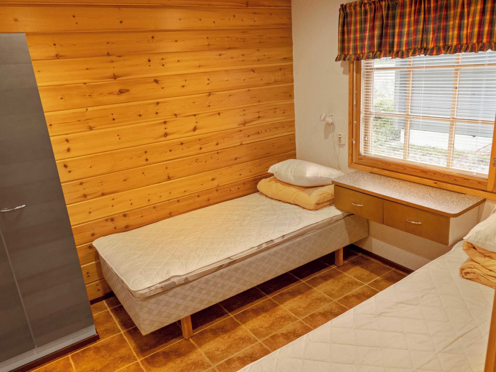 A small, single-bed room with a wooden wall, a window with blinds, a bedside table, and a wardrobe.
