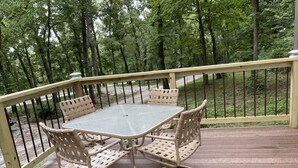 A wooden deck with a table and chairs, surrounded by a metal railing and a forested area.