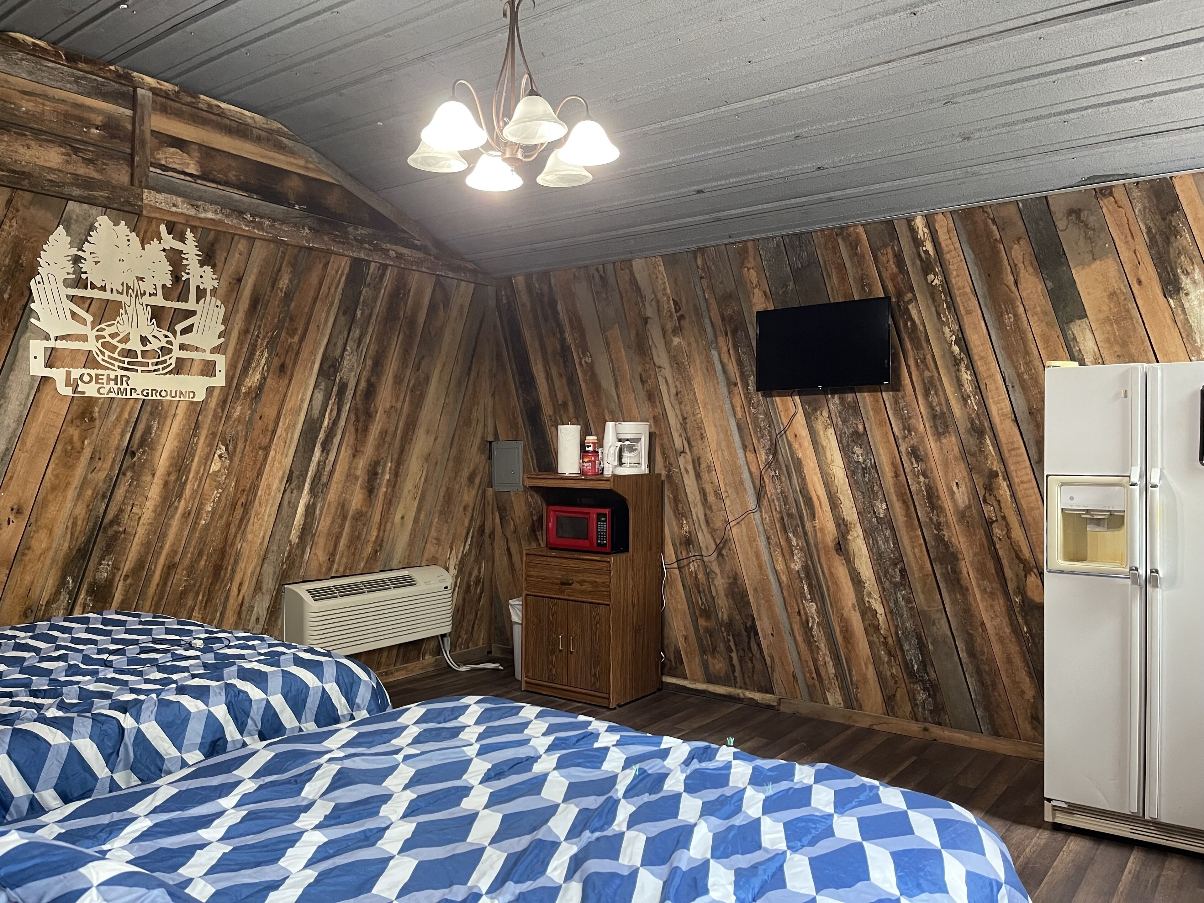 A room with a wooden slat wall, a bed with blue and white checkered bedding, a white refrigerator, and a mounted television.