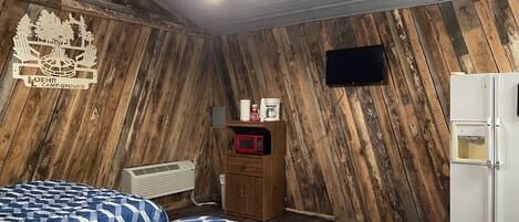 A room with a wooden slat wall, a bed with blue and white checkered bedding, a white refrigerator, and a mounted television.