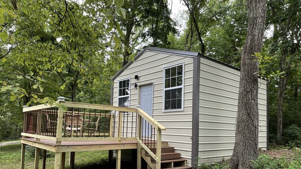 A small wooden cabin with a deck and a door, surrounded by trees.