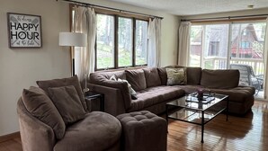 A spacious living room with a large sectional sofa, a glass coffee table, and a view of the outdoors through large windows. - Fire Pit, Babbling Brook, Poker & Foosball at this 3br+ home (Lakeville)