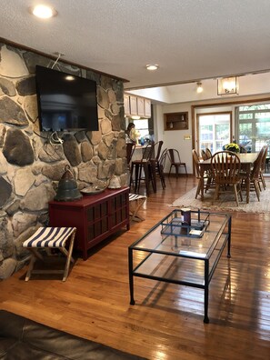 A spacious living room with a stone wall, a flat-screen TV, a wooden dining table with chairs, and a glass coffee table.