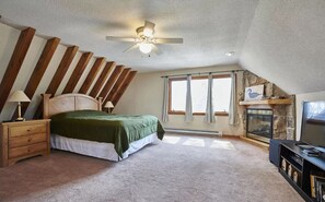 A bedroom with a sloped ceiling, a bed with a green bedspread, a nightstand with a lamp, a fireplace, and a TV stand with books.