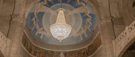 A grand hall with a large, ornate chandelier hanging from the ceiling, surrounded by detailed frescoes and classical architecture.