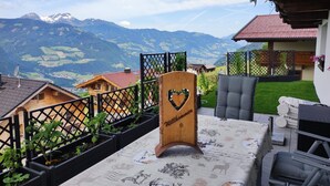 Une enseigne en bois avec l’inscription « Willkommen » et un cœur, une terrasse avec une table et des chaises, et un paysage montagneux en arrière-plan.