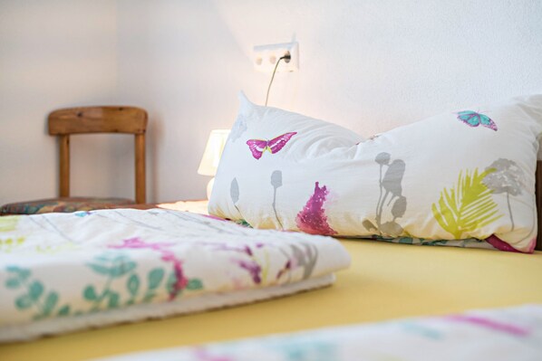 Un lit recouvert d’une couvre-lit à motifs floraux, une chaise en bois et une prise électrique murale.